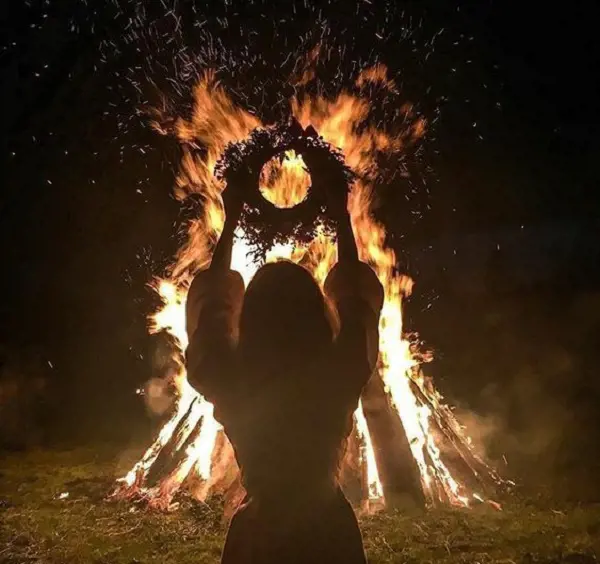A woman holds up a wreath in front of a huge bonfire to traditionally celebrate Samhain.
