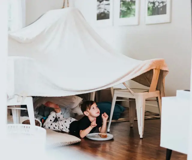 A child lays under an indoor fort made from bedsheet and enjoys his snack