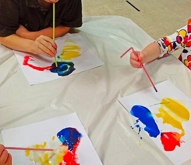 Children gather around a table to paint with primary color straws 