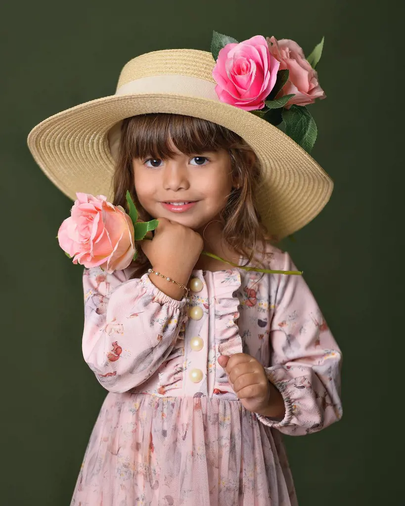 A studio shot captures a little girl in a dress and hat holding a rose flower