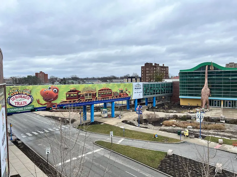 A sky bridge holds up Dinosaur Train sign while two Dinos are seen crawling inside Children's Museum Indianapolis' building. 